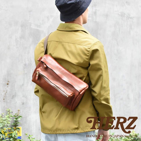 一款棕色皮革製成、帶有雙拉鍊設計的兩用腰掛斜背包，由一位穿著黃綠色外套和黑色帽子的人斜背著。