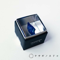 深藍色盒裝內，盛裝著一個金屬立方體，內部有一塊不規則的深藍色青金石原礦，表面帶有金色斑點。