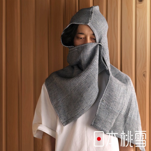 一位男子頭戴連帽浴巾，浴巾為灰白格紋，覆蓋住臉部，僅露出眼睛，背景為木質牆面。