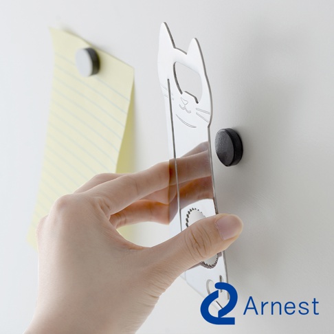 銀色金屬材質的貓咪造型開罐器，帶有貓咪臉部線條雕刻，可吸附在牆面，旁邊有一張黃色條紋紙和黑色磁鐵。