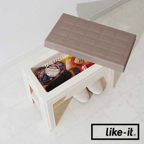 米白色塑膠製玄關穿鞋收納椅，上方置有棕色格紋椅蓋，內部收納有鞋油罐、鞋刷、鞋油瓶及一雙米色帆布鞋。