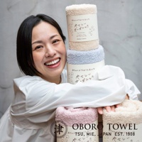 一位女士抱著三捲有機棉浴巾，分別為米白色、淺藍色和粉紅色。浴巾材質柔軟，捲起後呈現蓬鬆感，包裝上有日文標示。