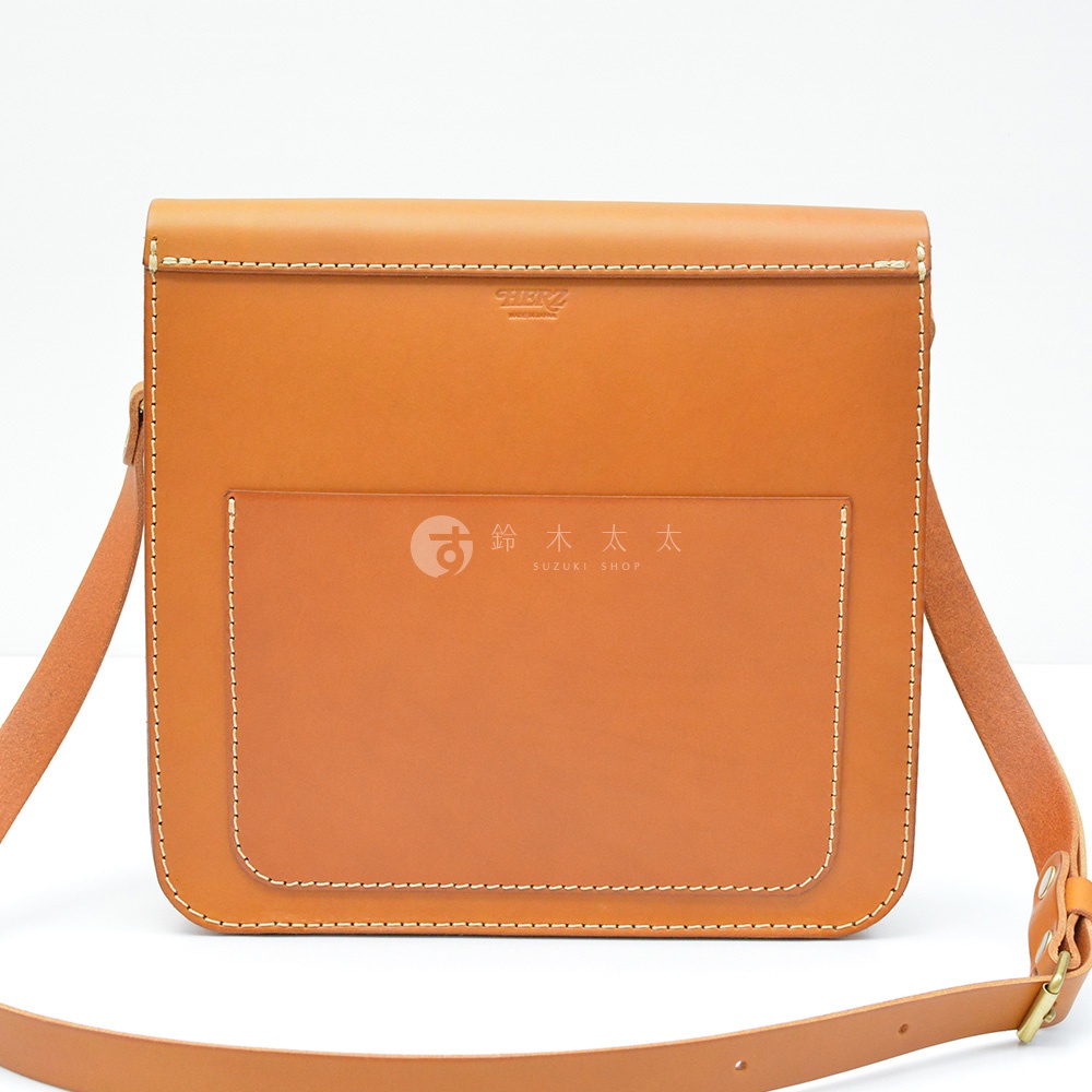 淺棕色皮革製作的厚實單肩包，正面有方形貼袋，袋蓋上印有品牌標誌。包包線條簡潔，縫線清晰，配有可調節的皮革肩帶。