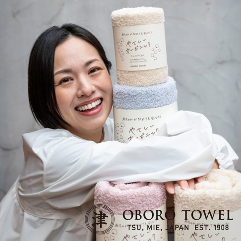 一位女士抱著三捲有機棉浴巾，分別為米白色、淺藍色和粉紅色。浴巾材質柔軟，捲起後呈現蓬鬆感，包裝上有日文標示。