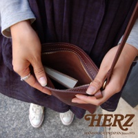 職人手作皮革Organ肩背小方包，棕色皮革，拉鍊開合，內部可見一本白色書本，由雙手持握。