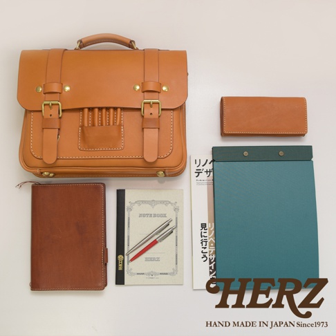 職人手作皮革傳統歐風三用學生包，淺棕色，配有兩個金屬扣環和提把。旁有同色系長夾、筆記本、鋼筆與筆記板。