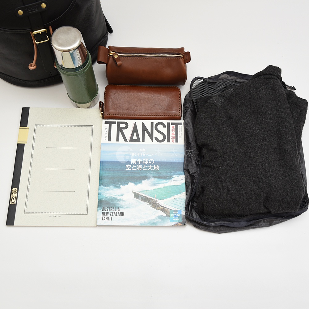黑色皮革束口後背包，搭配綠色保溫瓶、棕色皮革長夾、棕色皮革筆袋、雜誌和黑色摺疊收納袋。