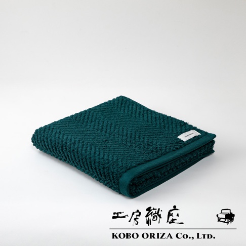 一條深綠色、觸感柔軟的有機棉浴巾，表面織有明顯的人字紋路，角落縫有白色品牌標籤。