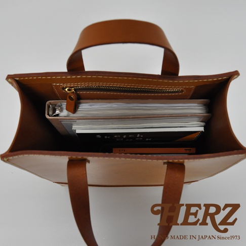 職人手作皮革直式托特包，淺棕色皮革，包內裝有筆記本和畫冊，頂部有拉鍊隔層和提把。