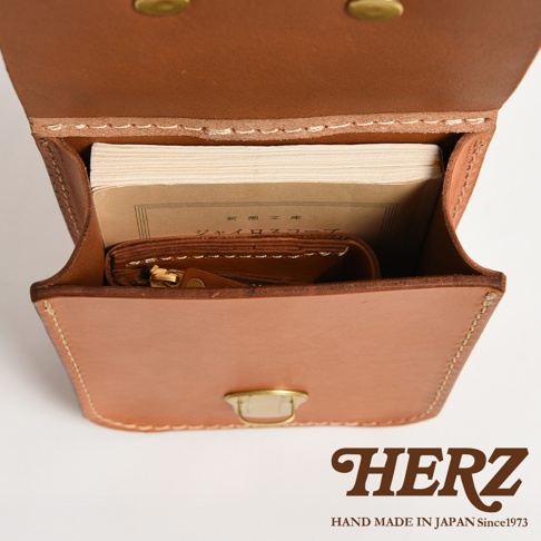 職人手作皮革經典腰袋E-49，淺棕色皮革，袋內裝有書籍和小錢包，整體線條簡潔，搭配黃銅釦件。