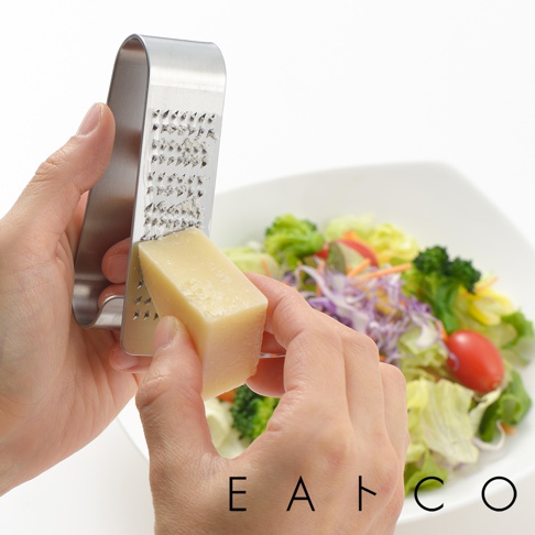 雙手持用不鏽鋼立式磨泥器，將一塊淺黃色起司磨碎，背景為一碗色彩繽紛的沙拉。