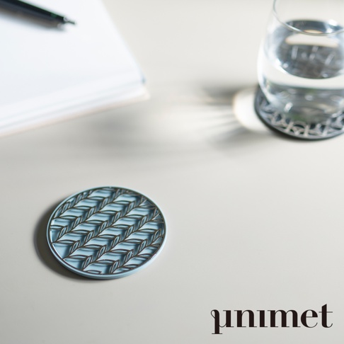 這是一張以淺色桌面為背景的產品情境照。桌面上放置著一個 Unimet 不鏽鋼光影藝術防滑杯墊，其上方鏤空呈現出精緻的葉脈紋路。右上方有一個盛水的透明玻璃杯，下方同樣墊著一個相同的杯墊。畫面左上角隱約可見紙張與筆，整體視覺風格簡約且具有現代設計感，右下角標註了品牌名稱 Unimet。
