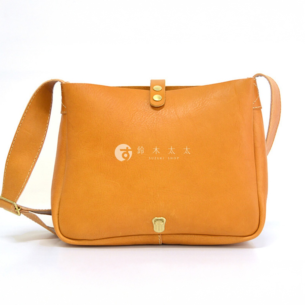一個琥珀色皮革橫式斜背包，正面有雙釦設計和一個金屬旋轉鎖扣，左側印有「鈴木太太 SUZUKI SHOP」的日文標誌。