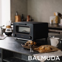 黑色 BALMUDA 蒸氣烤麵包機，配有金色邊飾，前方有玻璃門和兩個旋鈕。烤麵包機擺放在有咖啡器具和烤麵包的廚房檯面上。