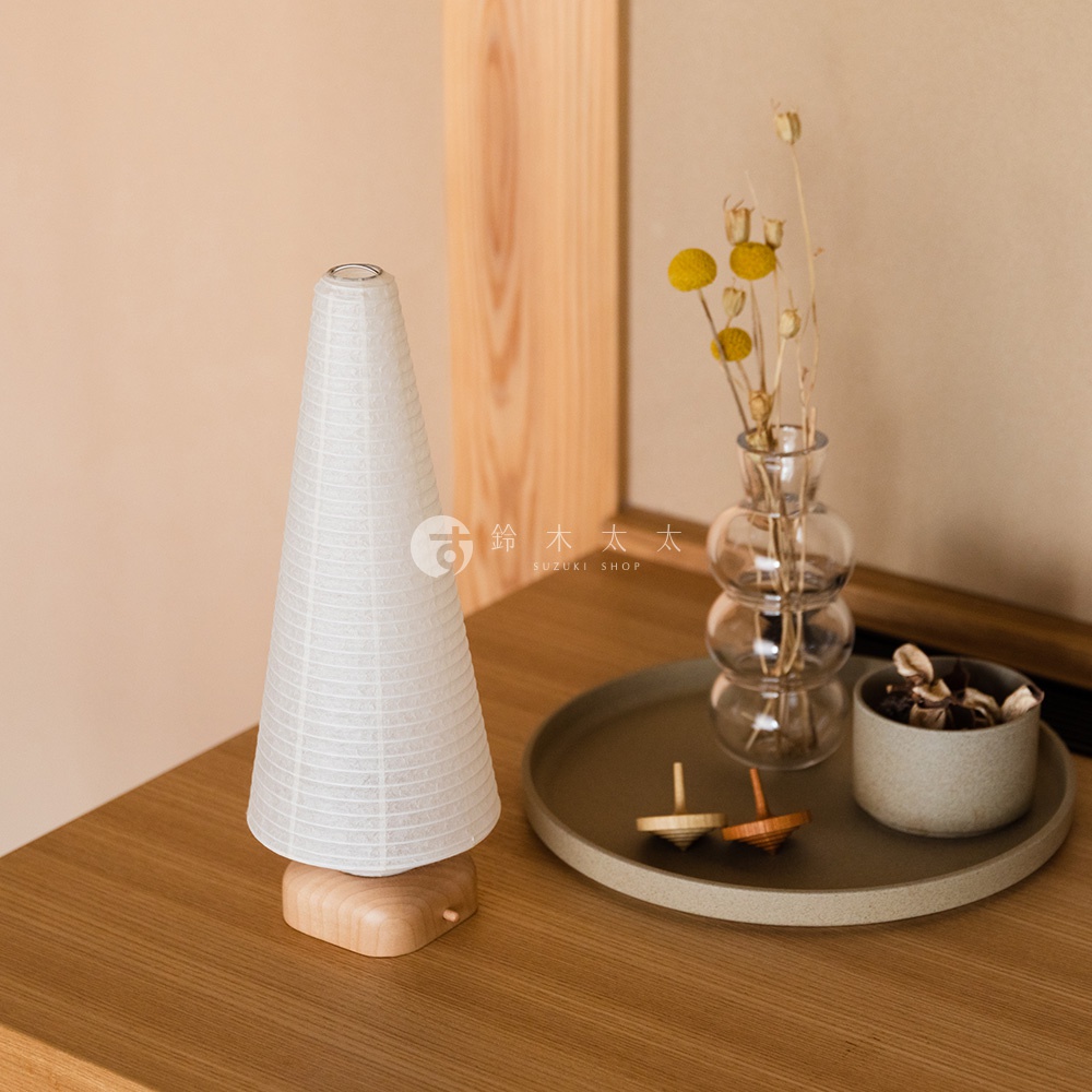 一個錐形白色紙質提燈，帶有木質底座，放在木桌上。旁邊有一個圓形托盤，上面擺放著兩個木製陀螺、一個裝有乾花的玻璃花瓶和一個裝有乾花的陶瓷碗。