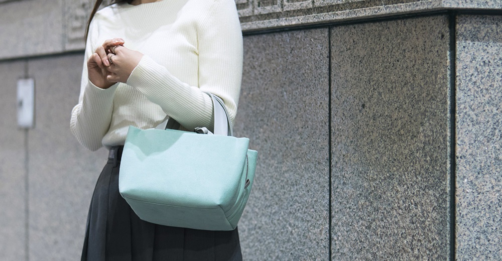 一名女子手持淺藍色托特包，身穿白色高領毛衣和深灰色長裙，背景為灰色斑駁牆面