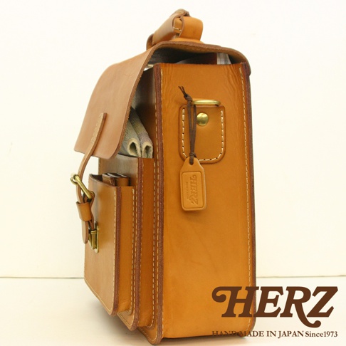 職人手作皮革單肩兩用小方包，採用天然棕色皮革，側面展示精緻縫線與金屬扣環，右側掛有品牌皮製吊牌。