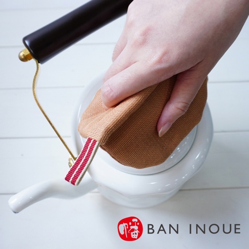 手持淺棕色、蚊帳質地的防燙鍋套，鍋套上有紅白色條紋織帶，正套在白色水壺的壺身上。