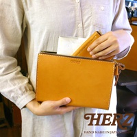 職人手作皮革三角底拉鍊手拿包，以天然皮革製成，呈現溫潤的焦糖色。簡潔的長方形設計，搭配同色系的車縫線和金屬拉鍊，展現細緻工藝。手拿包內有一小皮夾，方便收納。