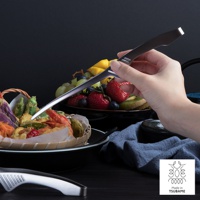 手持銀色不鏽鋼公筷夾，夾取籃中炸物。背景為一盤色彩鮮豔的水果，包含草莓、奇異果、葡萄和芒果。