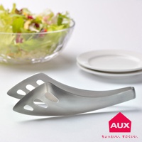 不沾桌沙拉料理夾，採用不鏽鋼材質，呈現銀色金屬光澤，夾子前端有橢圓形孔洞設計，方便瀝水。背景為一碗盛滿新鮮沙拉的玻璃碗及疊放的白色盤子。