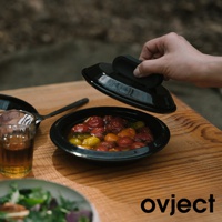 一隻手正拿起一個黑色琺瑯鍋的鍋蓋，鍋內裝有豐富的番茄和香草，旁邊擺放著一個裝有飲料的玻璃杯和餐具，桌面為木質。