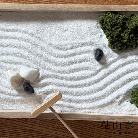 一個長方形的木製日本枯山水模型，細緻的白色沙面上耙出了流暢的波浪紋理，沙中點綴著幾顆黑色與白色的圓潤鵝卵石，角落處有青苔裝飾，下方放置著一把木製耙子，右下角印有枯山水及英文拼音 karesansui 字樣。