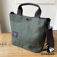 軍綠色帆布材質的兩用托特包，配有黑色皮革提把和可拆卸肩帶，包身正面有紅色標籤，標有日文文字和黑犬圖案。