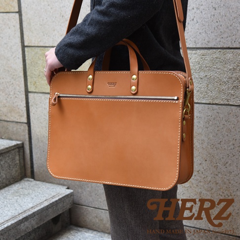 職人手作皮革兩用商務公事包，採用鞣製皮革製成，呈現自然棕色調。包款設計簡潔，帶有頂部提把及可拆卸肩背帶，適合商務場合使用。包身上有品牌標誌及精緻車縫線。