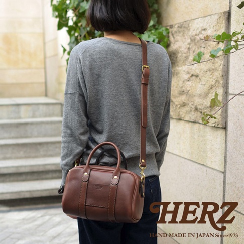 日本HERZ職人手作皮革兩用迷你波士頓包，模特背著棕色小牛皮包，包款有手提把和可拆卸肩帶，包身上印有HERZ品牌標誌。