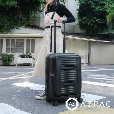 一位穿著黑色上衣與米白色吊帶裙的女性，正站立在戶外街道上，手扶著一個霧面黑色的AZPAC品牌行李箱，該行李箱採用前開口設計並配備伸縮拉桿與多向輪。