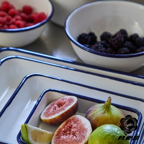 白色琺瑯調理盤，邊緣飾有藍色條紋，盤中盛有切半的無花果、檸檬塊和一顆青檸。背景為裝有覆盆子和黑莓的琺瑯碗。