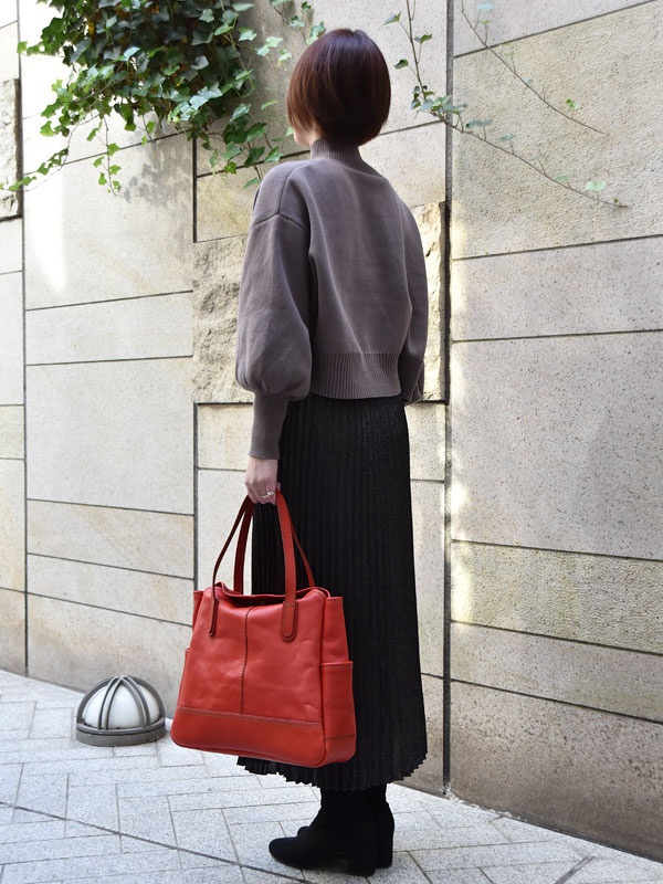 紅色皮革直式托特包，包身縫線明顯，兩側有插袋，搭配棕色短袖上衣和黑色長裙。