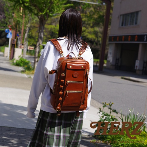 棕色皮革製成的職人手作兩用後背包，具有多個拉鍊口袋和金屬扣環，背在穿著白色上衣和格紋裙子的模特兒身上，於戶外行走。