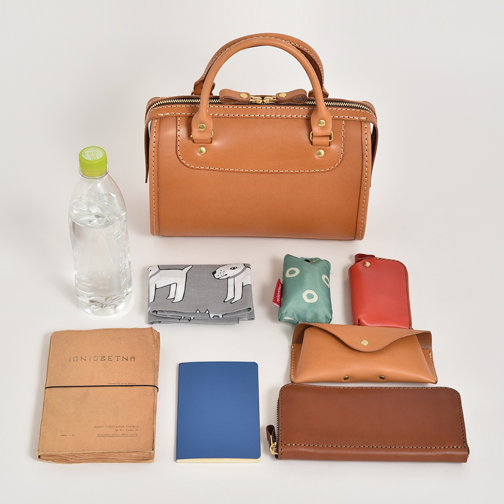 職人手作皮革迷你波士頓包，淺棕色，附有頂部提把、拉鍊開合，以及金屬扣飾。周圍擺放著一瓶水、印有狗圖案的灰色布料、綠色小包、紅色鑰匙包、淺棕色皮質眼鏡盒，以及兩本筆記本（一本深棕色，一本藍色）。