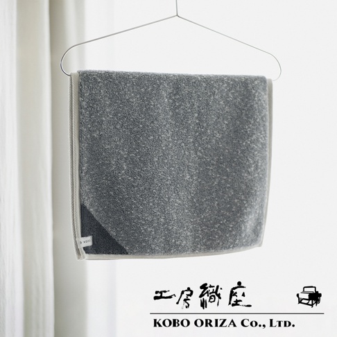 灰色棉麻混紡雙色快乾浴墊，邊緣帶有淺灰色滾邊，懸掛在衣架上。