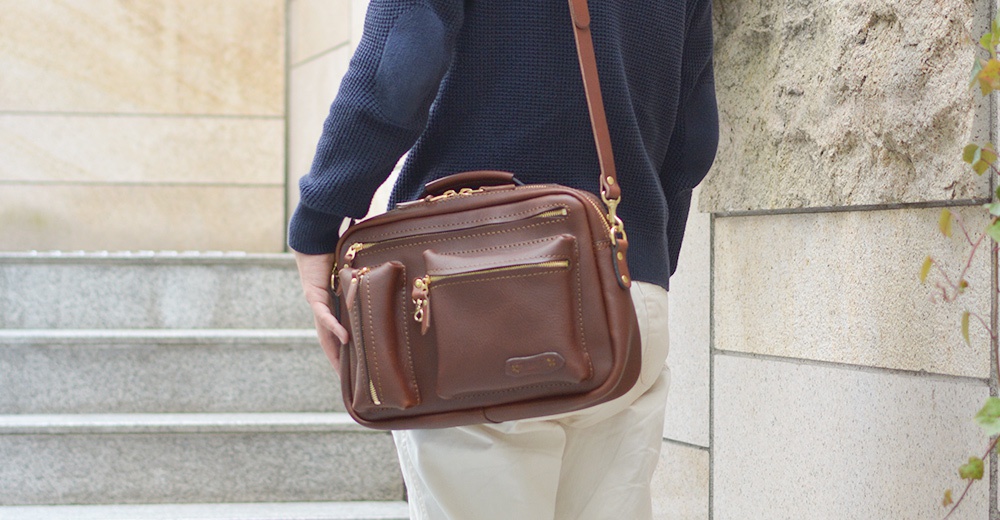 咖啡色皮革製成的手提/斜背兩用包，有多個拉鍊口袋及一個旋釦前袋，搭配金色五金配件，展現職人手作質感。