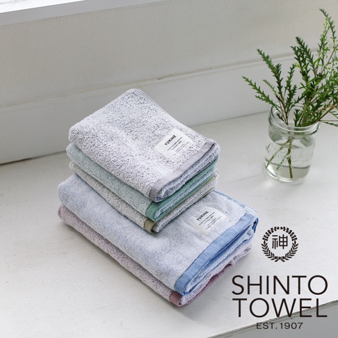 一疊YUKINE雪中漫步有機棉蓬厚毛巾，顏色包含淺灰、薄荷綠、淺藍及粉紫，毛巾表面蓬鬆，帶有精緻的織紋，邊緣有不同顏色的織邊，右側有綠色植物裝飾。