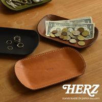 職人手作皮革小物收納盤，共有黑、棕、綠三色。圖片展示了黑色收納盤內有戒指，棕色收納盤內有鈔票和硬幣，以及一個未裝物品的淺棕色皮革收納盤，盤上壓印有「HERZ」字樣。
