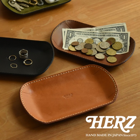 職人手作皮革小物收納盤，共有黑、棕、綠三色。圖片展示了黑色收納盤內有戒指，棕色收納盤內有鈔票和硬幣，以及一個未裝物品的淺棕色皮革收納盤，盤上壓印有「HERZ」字樣。