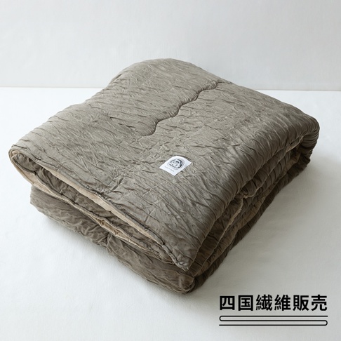 一張摺疊起來的單人被放在白色背景上，被面呈現灰棕色的皺褶紋理，觸感看似柔軟。被子右側中央有一個寫著日文的白色布質標籤，右下角印有四國纖維販賣的黑色字樣，整體呈現簡約的日式家居風格。
