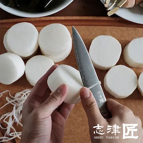 一雙手正使用一把有著達馬斯克紋路的銀色金屬主廚刀，削切著圓形的白色蘿蔔塊。刀柄上有日文標記，背景是木製砧板和一碗深色食材。