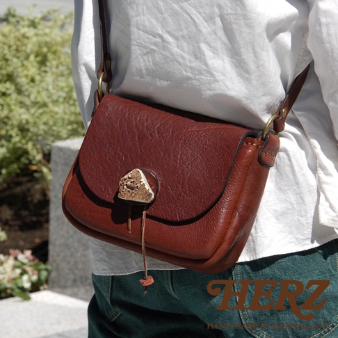 棕色皮革製作的職人手作肩背包，包蓋處飾有鹿角造型的掛扣及一條細繩，包款為斜背包設計，搭配金屬環扣與皮革背帶。