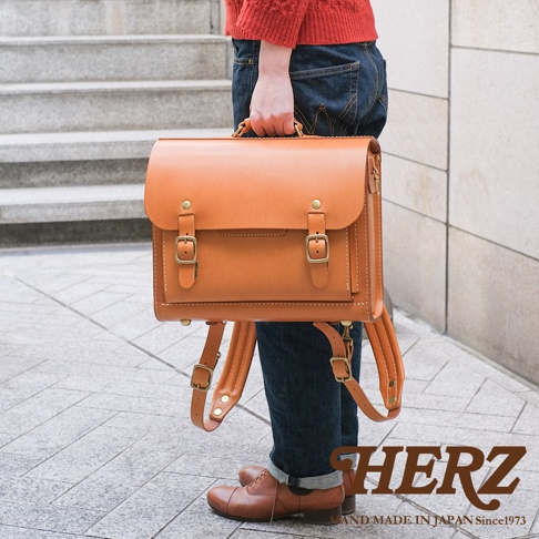 一位身穿紅色毛衣和牛仔褲的人，手提一個棕色皮革製的橫式書包，書包上有金屬釦帶和縫線細節，腳穿棕色皮鞋。