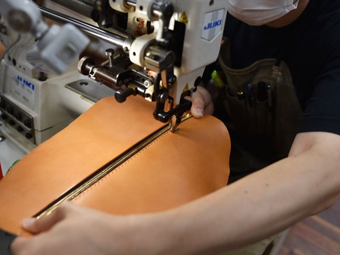 職人正在使用工業縫紉機縫製一個淺棕色皮革製的立體斜背包。皮革表面光滑，縫線清晰，拉鍊為金色。