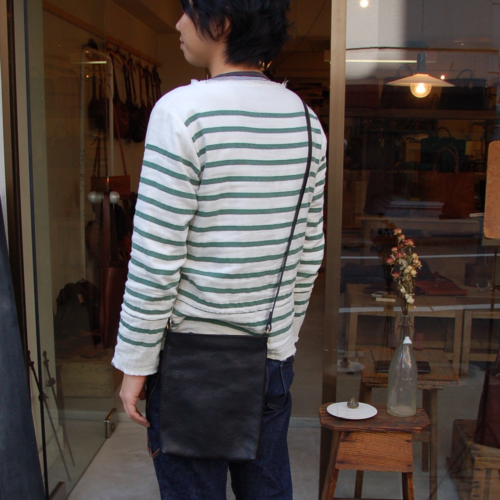 一位男性身著綠白條紋長袖上衣，背著一個黑色皮革製的肩背包。包款為方形設計，質感柔軟，背帶細長，跨掛於右肩。