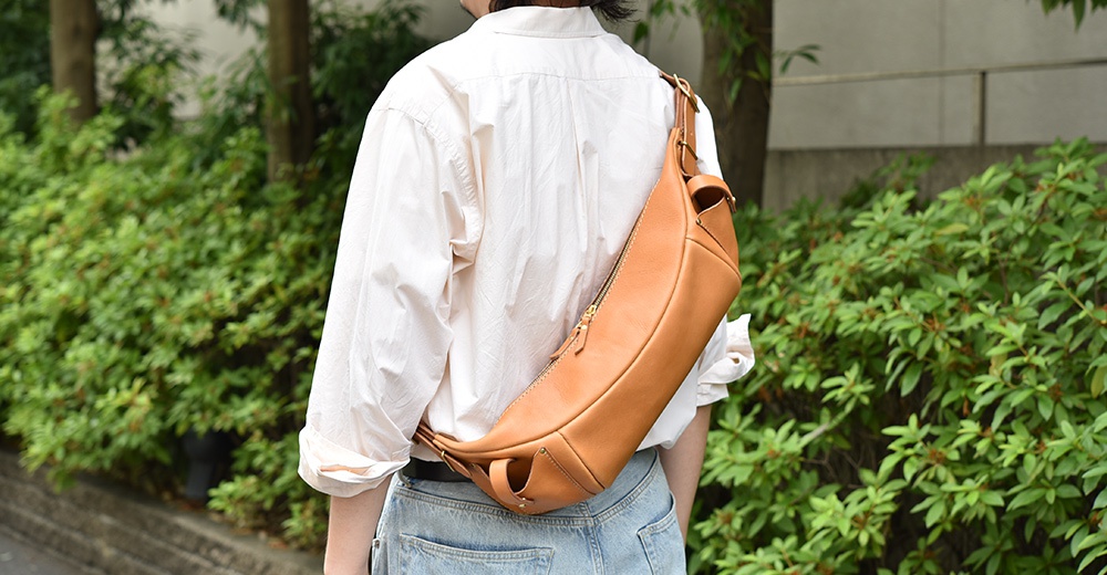 日本HERZ職人手作Stare皮革大容量斜背包，棕色皮革，肩背帶搭配銀色金屬拉鍊，背面視角，背景為綠色植物。
