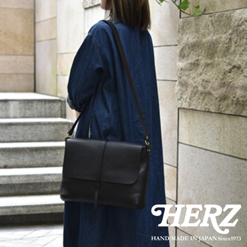 一位身穿藍色長袍的女士，斜挎著一個黑色的皮革馬鞍包，包上配有細扣帶，包的下方壓印著 HERZ 的 Logo 和 HAND MADE IN JAPAN Since1973 的字樣。