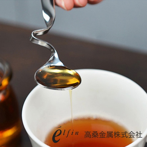 日製不鏽鋼製的螺旋醬料匙，匙面盛著琥珀色蜂蜜，正滴入白色杯子內的茶飲。