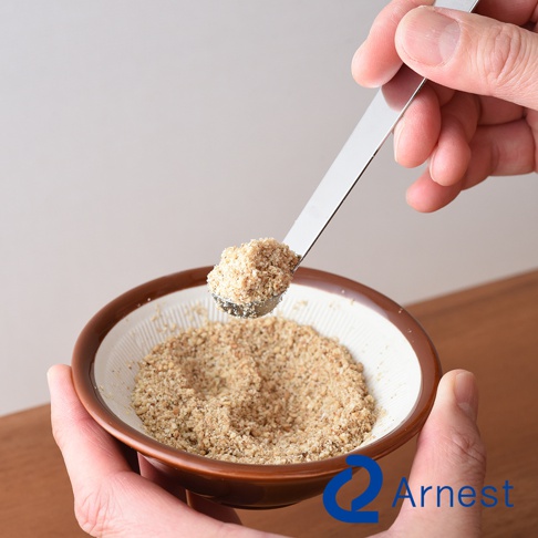 手持不鏽鋼小勺，舀取陶瓷臼中研磨的淺棕色粉末。小勺為銀色金屬材質，勺頭裝有少量細碎粉末。